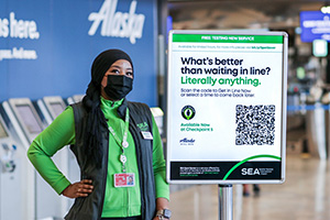 Airport staff member beside a sign promoting a free queuing service, with self-service kiosks in the background.