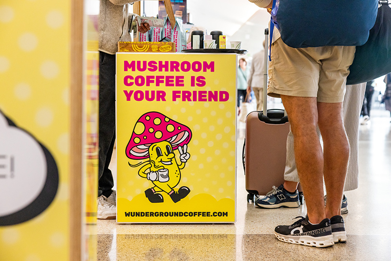 Sign at a Wunderground coffee kiosk that reads 'Mushroom coffee is your friend,' with travelers standing nearby.
