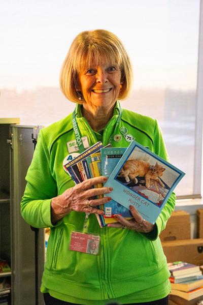 Becky Nickels wearing a green SEA Airport volunteer jacket holds several books and magazines in front of a small shelving area.