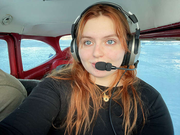 Elise Dempster wearing a headset sits inside a small aircraft with windows showing a snowy landscape outside.