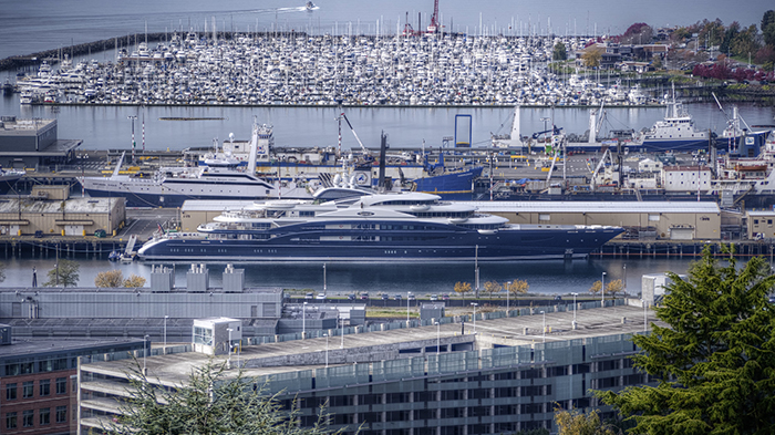 A large yacht docked in an industrial harbor with shipyards, cranes, and a dense marina of boats in the background.
