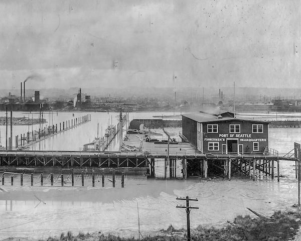 A historic black‑and‑white photo of Fishermen’s Terminal showing the Port of Seattle Fishermen’s Headquarters building on a pier, with docks, moored vessels, and an industrial waterfront visible in the background.