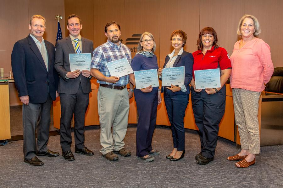 Group of people standing in a conference room holding certificates, with a backdrop showing a podium and a flag. The individuals are positioned in a row and appear to be part of an awards or recognition event.