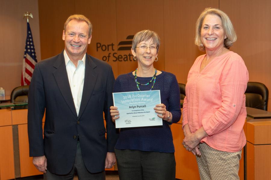Three people standing in a conference room, with the person in the center holding a certificate for the 2018 2nd Quarter Innovation Award on behalf of the Honorable Mention recipients. The background includes a Port of Seattle sign and an American flag.