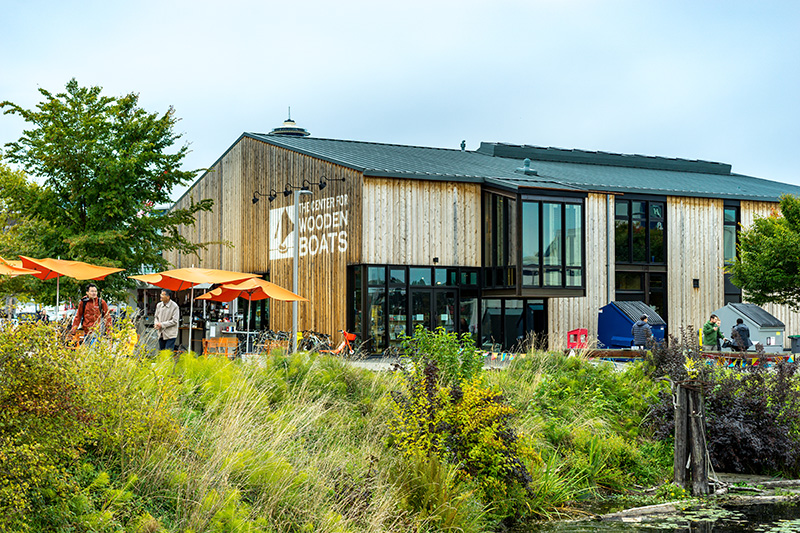 The exterior of the Center for Wooden Boats, a modern wooden building with large windows, surrounded by greenery and outdoor seating with orange umbrellas.