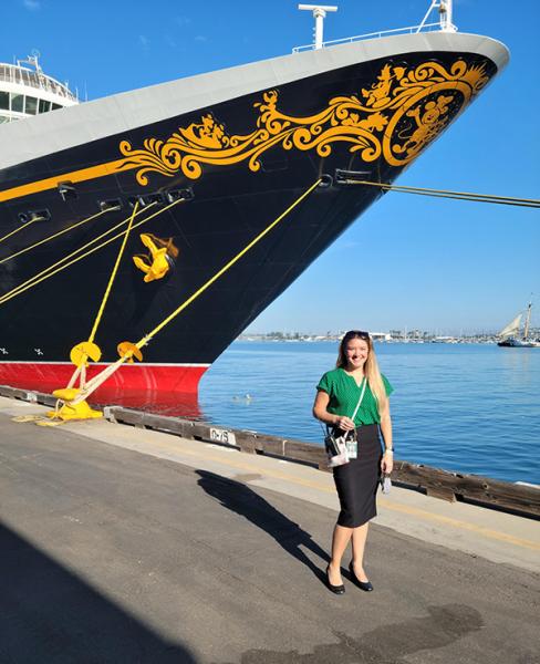 A person stands on a pier in front of a large ship with ornate gold detailing near the bow on a clear day.