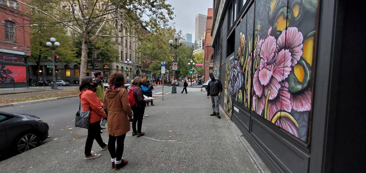 A small group of people gathers on a downtown sidewalk near a building decorated with a large, colorful floral mural. The street is lined with older brick buildings, trees, and traffic signals, with a few pedestrians and cars visible in the background.
