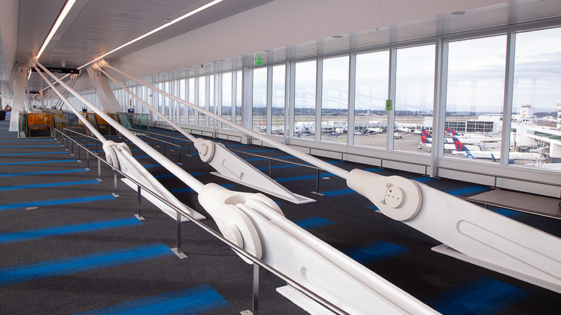 “A bright airport walkway with large floor‑to‑ceiling windows overlooking aircraft parked on the tarmac. The corridor features blue‑striped carpet and prominent white structural cables anchored along the walkway, with handrails running beside them.