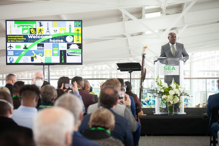 Speaker standing at a clear podium with a SEA Airport sign addresses an audience seated in an indoor event space. A large screen to the left displays graphics and the text 'International Arrivals Facility @ SEA Airport' and 'Welcome here. Welcome home.' The background shows tall windows and the airport interior. Floral arrangements sit at the base of the podium, and several audience members are taking photos.