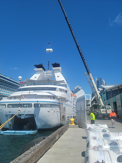 A cruise ship is docked at a pier while a crane lifts cargo overhead, with workers and equipment positioned along the waterfront.