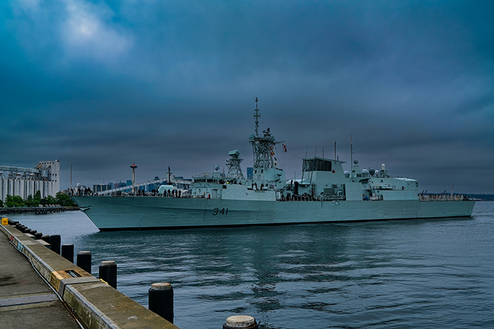 A naval ship with the number 341 moves through calm harbor waters under a dark, cloudy sky near a pier.