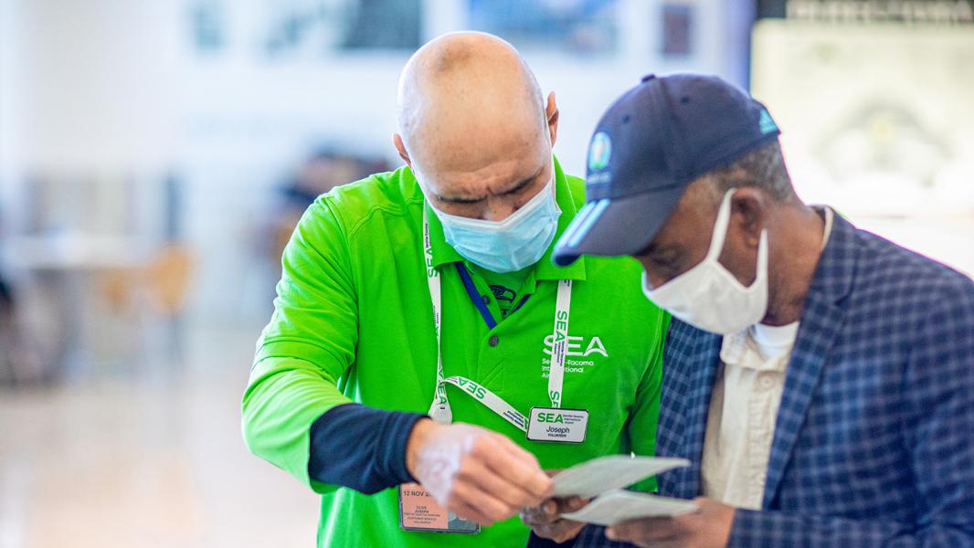 An airport staff member in a bright green shirt and face mask helps a traveler review documents, both leaning in to look closely.