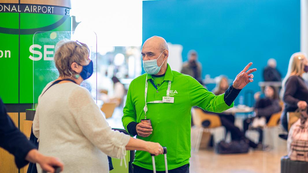 An airport staff member in a bright green shirt and face mask gestures while speaking with a traveler using a rolling suitcase near an information area.