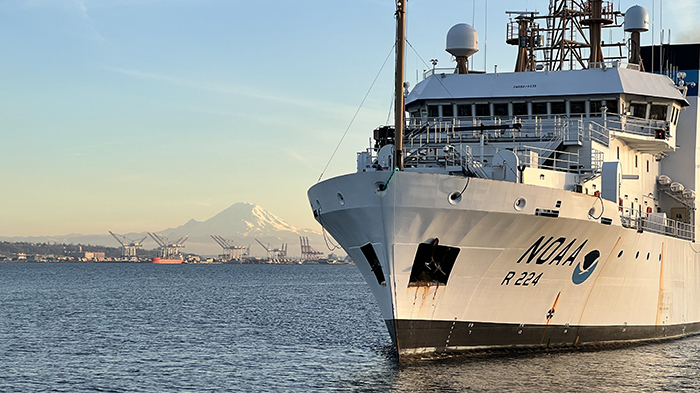 A large NOAA research vessel docked on calm water with a distant mountain and port cranes in the background.
