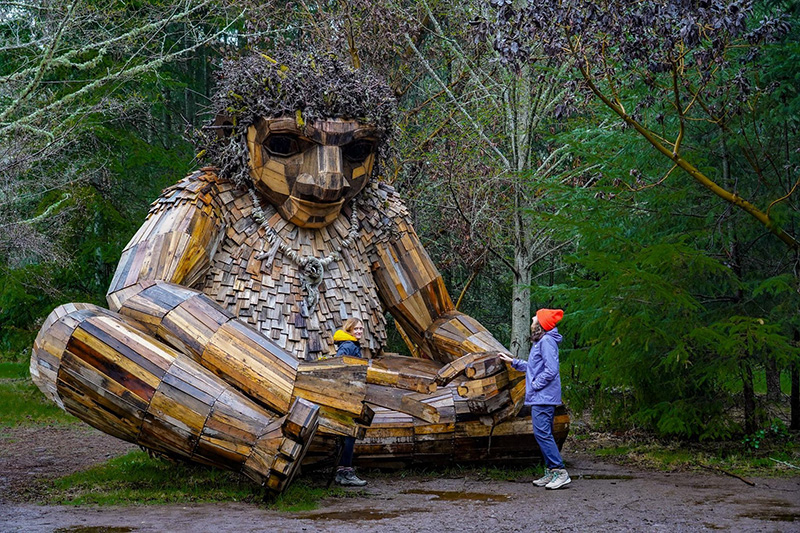 Pia the Peacekeeper Troll sculpture made of wooden pieces sitting in a forested area, with two people standing beside the large figure.