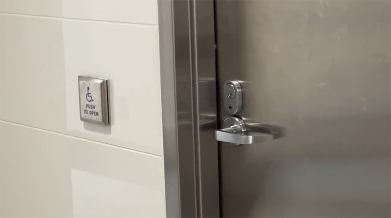 A close-up of a metal door with a lever handle next to a wall-mounted accessibility push-button labeled ‘Push to open.