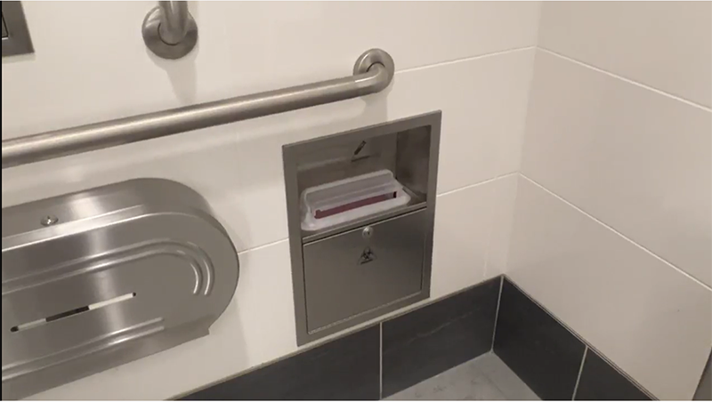 A restroom wall with a metal grab bar, a mounted toilet‑paper dispenser, and a recessed disposal unit with an open compartment.