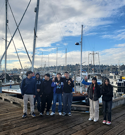 A group of people stand together on a wooden dock with numerous sailboats and masts behind them under a partly cloudy sky.