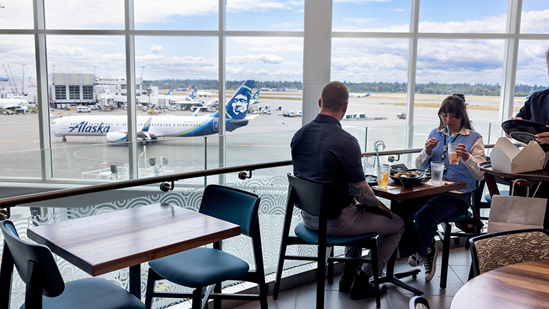 People sit at an airport restaurant with large windows overlooking Alaska Airlines planes on the tarmac.