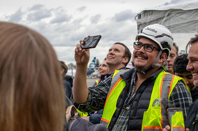 A group of people wearing safety vests and hard hats gather outdoors, with one person holding up a phone to capture a photo.