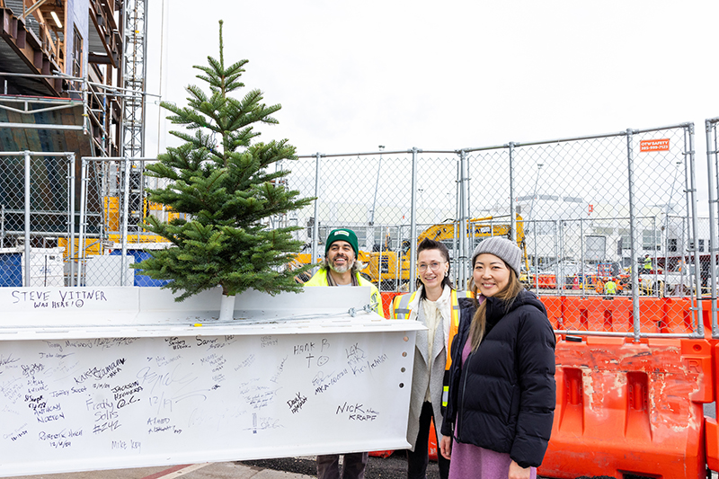 Three people stand beside a signed construction beam topped with a small evergreen tree at an active jobsite.