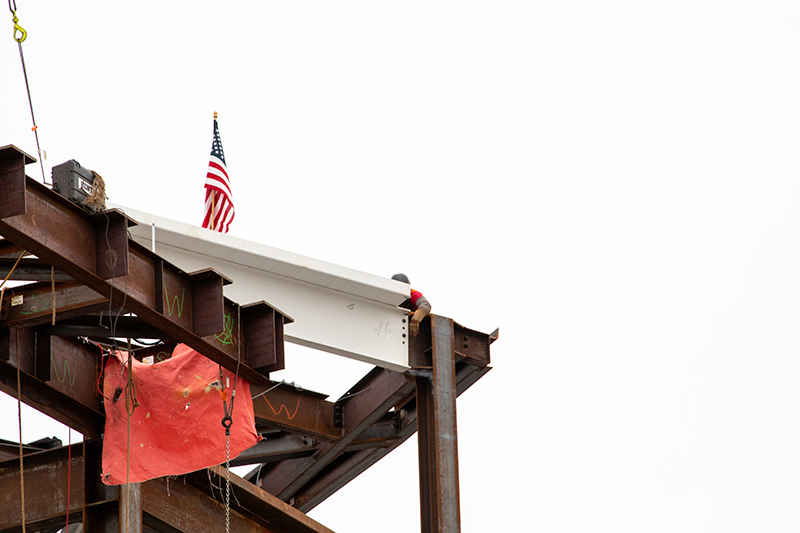 A construction worker stands on a steel structure as a signed white beam with an American flag is positioned at the top.