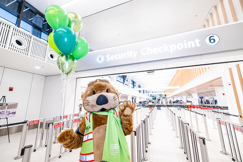 A costumed otter character named Jett stands in front of Security Checkpoint 6, holding a bunch of green balloons. The checkpoint area features bright lighting, metal stanchions, and a large overhead sign that reads “Security Checkpoint 6.”