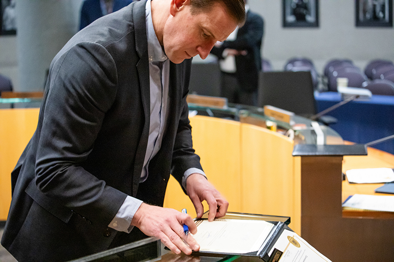 Commissioner Calkins in a suit leans over a desk in a meeting room, signing a document with a pen. The desk surface holds papers and a framed certificate, and curved wooden panels and chairs are visible in the background.