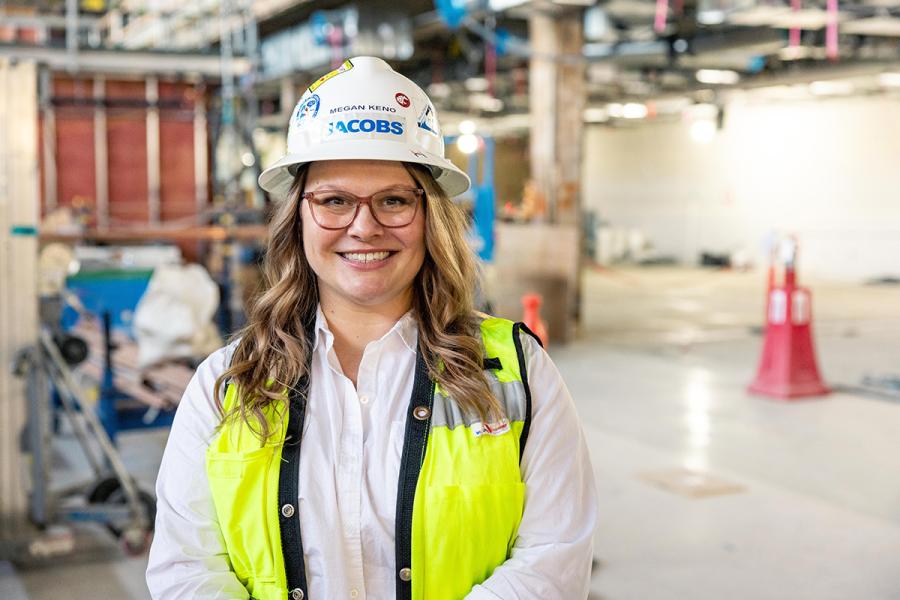 Megan Keno wearing a hard hat, glasses, and a bright safety vest stands inside an unfinished building with tools, materials, and construction equipment in the background.