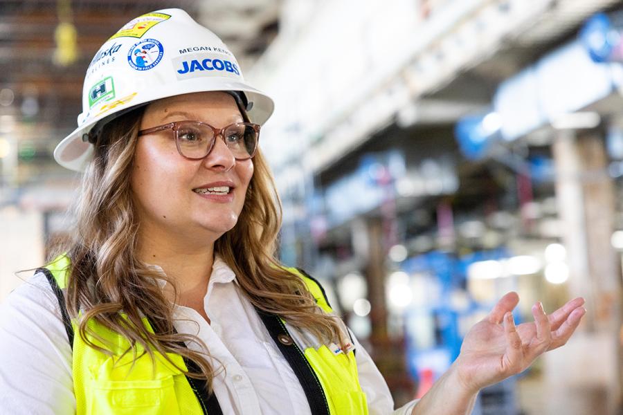 Megan Keno wearing a hard hat, glasses, and a high‑visibility vest speaks with one hand raised inside a busy construction area filled with overhead pipes and equipment.