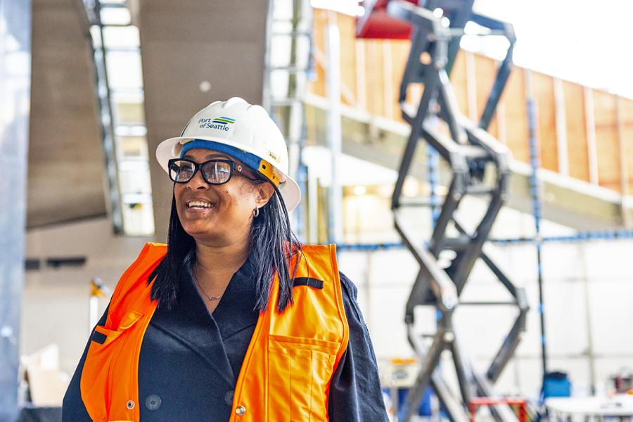 Tanisha Delgardo wearing a hard hat, glasses, and an orange safety vest stands at an indoor construction site with a scissor lift and unfinished structures behind them.