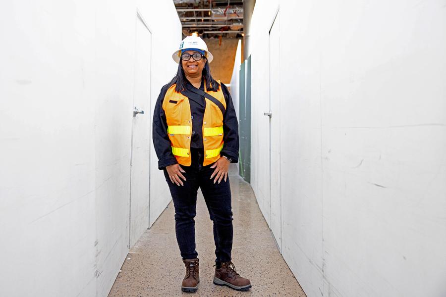 Tanisha Delgardo wearing a hard hat, glasses, and a high‑visibility vest stands in a narrow hallway with white temporary walls and an unfinished ceiling.