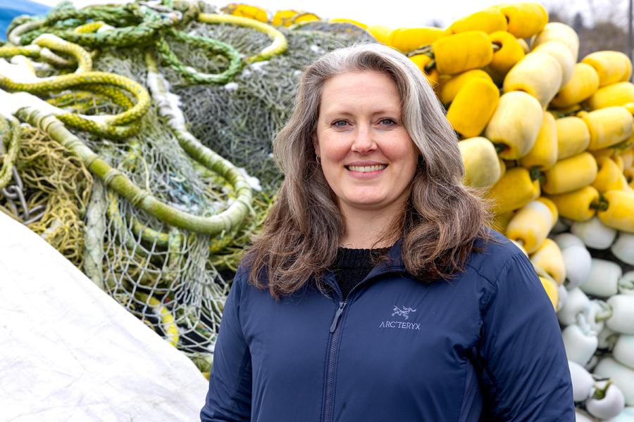 Kelly Purnell wearing a dark jacket stands outdoors in front of large piles of fishing nets and yellow and white floats.