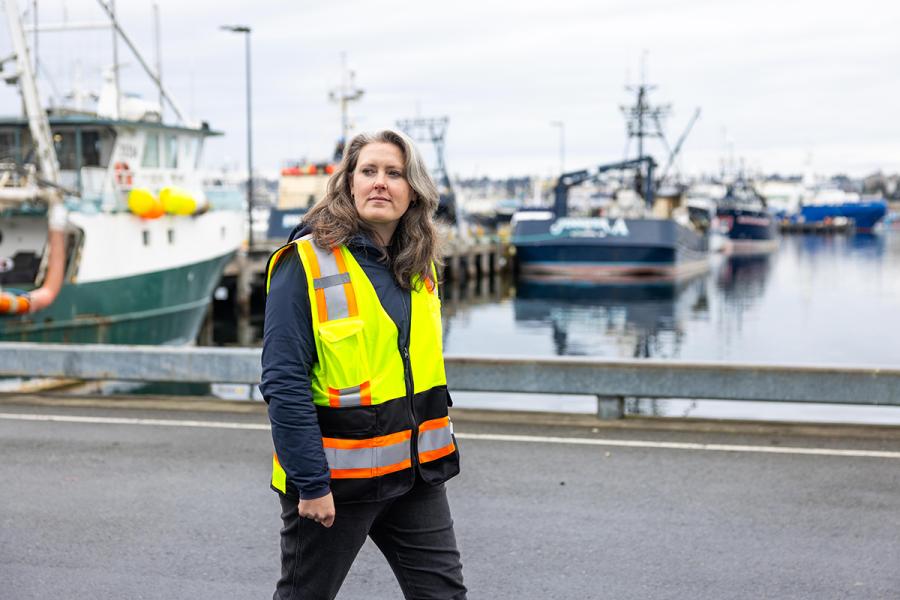 Kelly Purnell a high‑visibility safety vest walks along a waterfront area with several docked fishing vessels in the background.