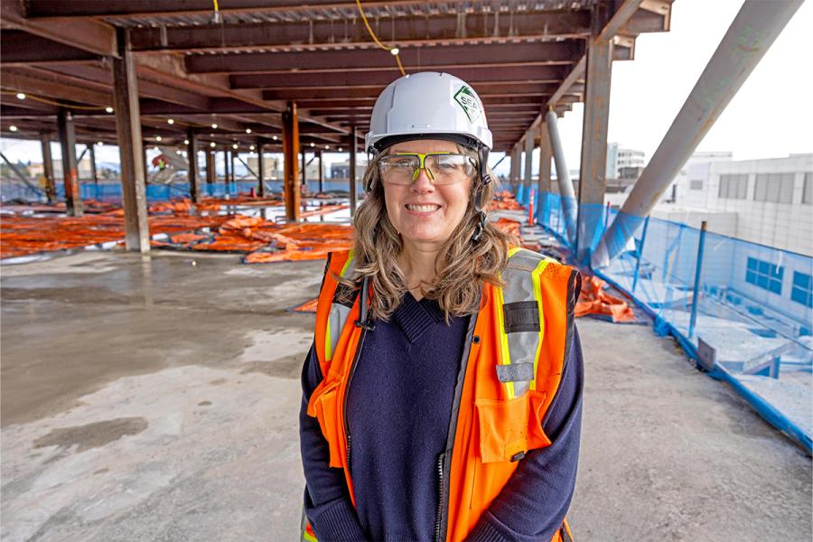Janet Sheerer wearing a hard hat, safety glasses, and an orange safety vest stands on an unfinished building floor with exposed beams, concrete surfaces, and blue safety netting in the background.