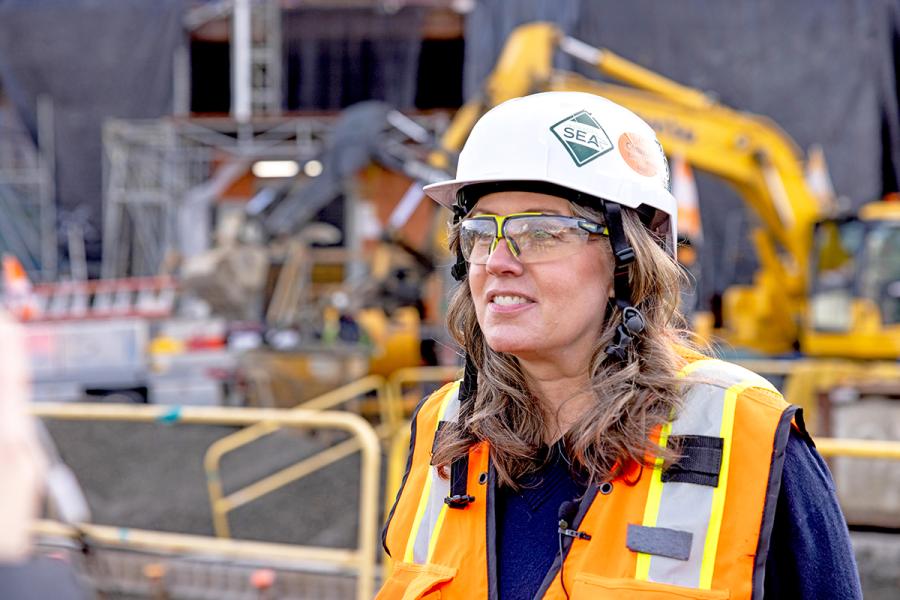 Janet Sheerer worker wearing a hard hat, safety glasses, and an orange safety vest stands at an active construction site with heavy equipment and scaffolding in the background.
