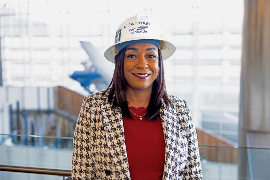 Lisa Phair wearing a Port of Seattle hard hat and patterned jacket stands in an indoor space with glass walls and railings in the background. The hard hat displays the text “Lisa Phair” and “Port of Seattle.