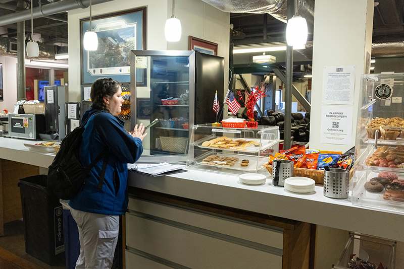 A person stands at a counter in a snack area with display cases holding pastries, crackers, and packaged chips. Small flags and signs are visible on the counter, along with hanging lights and framed items on the wall behind.
