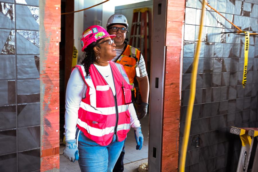 Two individuals wearing hard hats, safety vests, and work gloves walk through an indoor construction area with tiled walls, exposed framing, caution tape, and a ladder in the background.