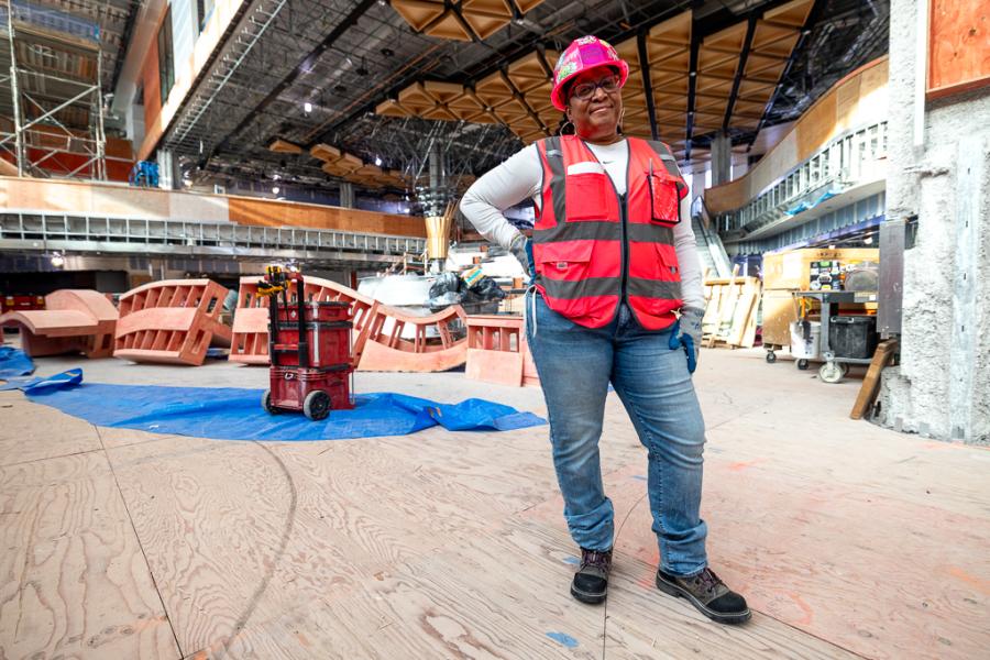 Nicolle Strong wearing a hard hat, safety vest, gloves, jeans, and work boots stands confidently in the middle of an active construction site filled with tools, wood structures, and scaffolding.