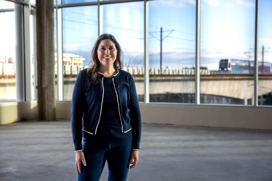 Regan Greenhill stands inside a large, empty building with tall windows, wearing dark clothing, with a rail line and train visible outside in the background.