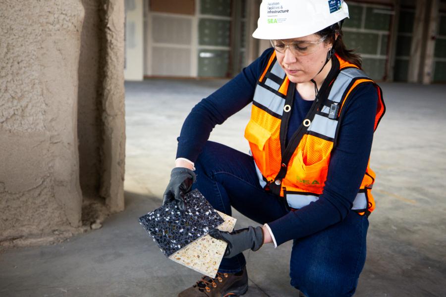 Regan Greenhill wearing a hard hat, safety vest, gloves, and protective eyewear kneels on a concrete floor in an unfinished building, examining two sample tiles—one dark and speckled, the other light and textured.