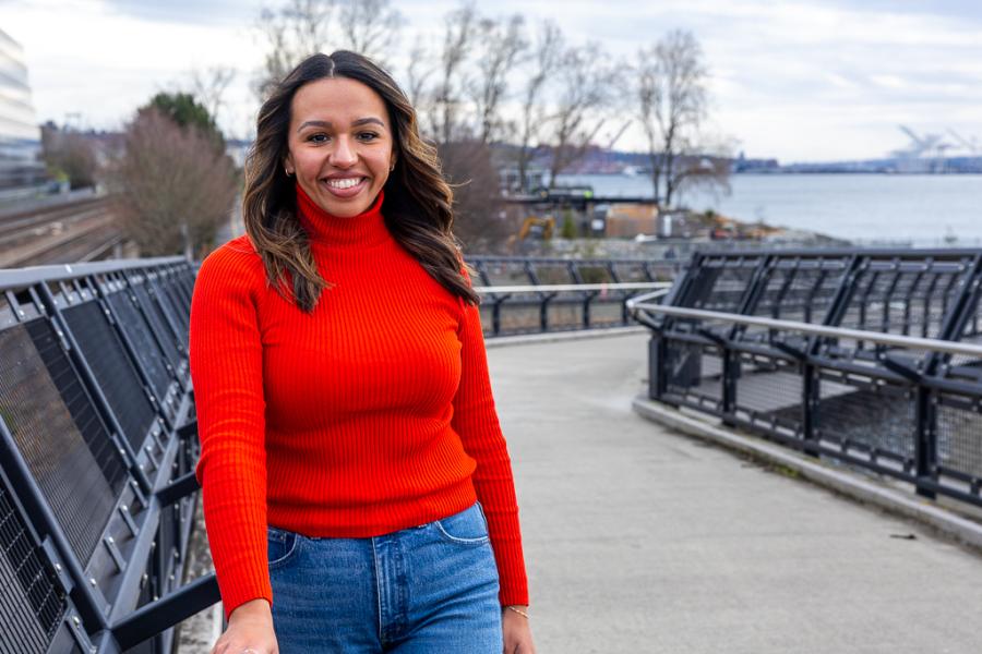 Crystal Cuellar walks along an outdoor pathway bordered by metal railings, with trees, railway tracks, and waterfront views in the background.
