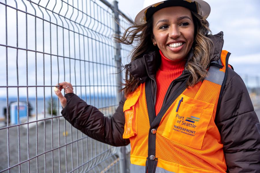 Crystal Cuellar wearing a hard hat, an orange safety vest labeled “Port of Seattle Waterfront Project Management,” and a dark jacket stands beside a metal fence at an outdoor work site, with water and equipment visible in the background.