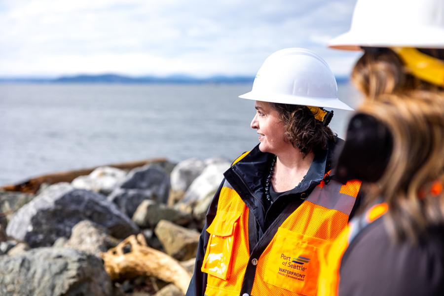 Two individuals wearing hard hats and orange safety vests stand on a rocky shoreline, looking out toward the water and distant land under a cloudy sky.