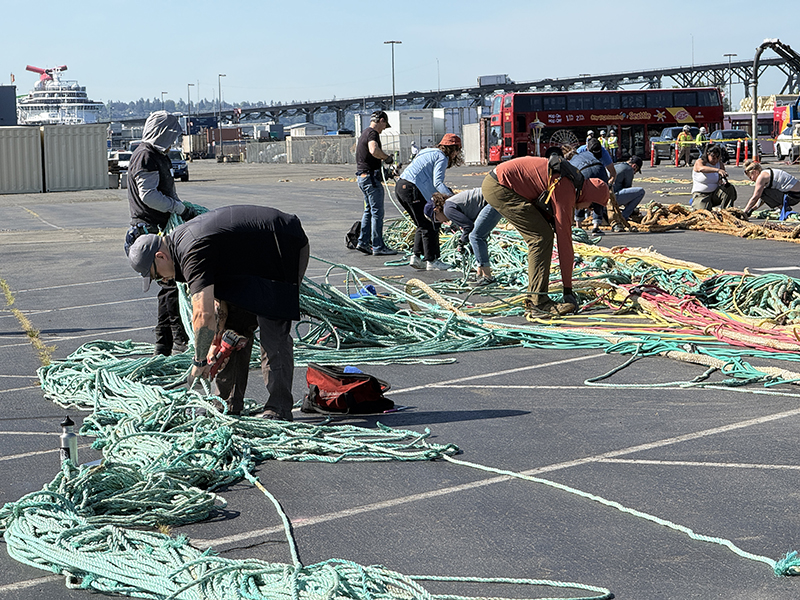 People are working in an outdoor lot, sorting and organizing long bundles of green and multicolored rope spread across the pavement. Storage containers, a double‑deck tour bus, and a large ship are visible in the background near a waterfront.