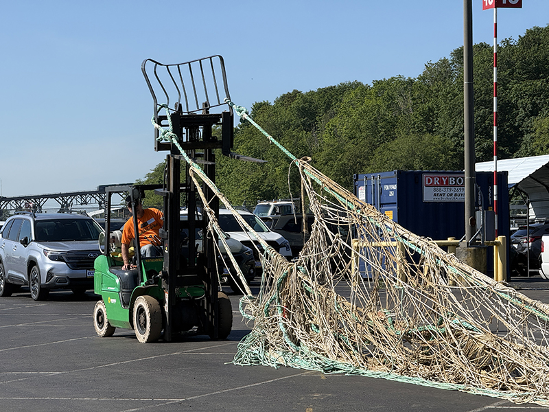 A forklift in an outdoor lot is dragging a large fishing net made of light‑colored rope across the pavement. Parked vehicles, shipping containers, and a tree‑covered hillside are visible in the background under clear skies.