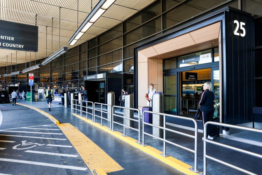 Exterior view of Door 25 in the airport departures area, showing the entrance, railings, and several people standing near the doorway, with vehicles and signage visible along the curbside.