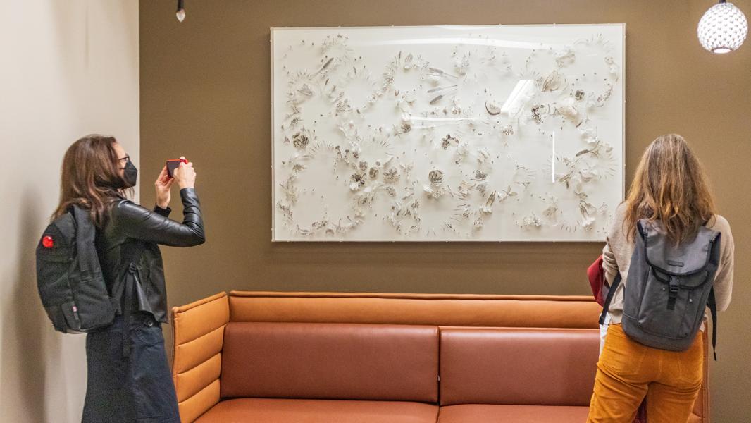 Two people viewing a textured white artwork mounted on a wall in a seating area.