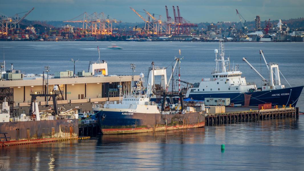 Commercial fishing vessels docked at an industrial waterfront pier, with cranes, shipping containers, and port infrastructure visible across the water in the background. The scene shows calm water and several large ships moored alongside buildings and equipment.